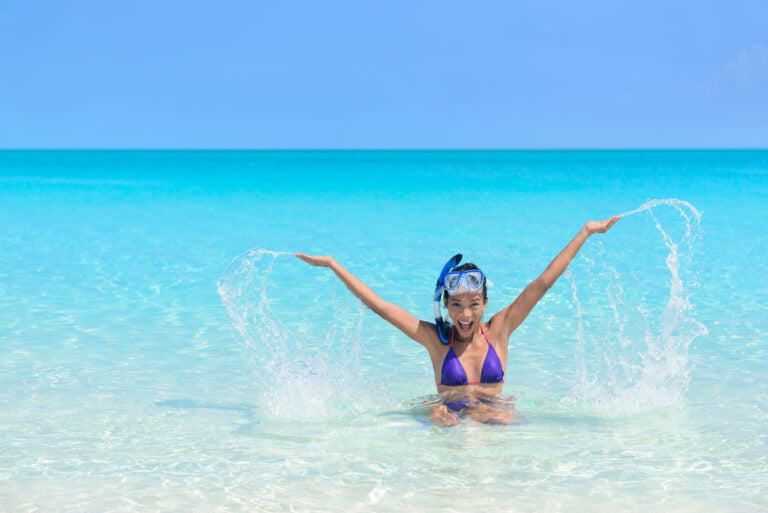 Beach holiday woman playing in the ocean. Asian young adult wearing a snorkel scuba mask having fun splashing water with arms up and swimming in vacation resort travel destination
